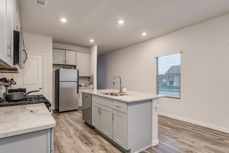 Kitchen with light wood-type flooring, light stone counters, sink, a center island with sink, and appliances with stainless steel finishes