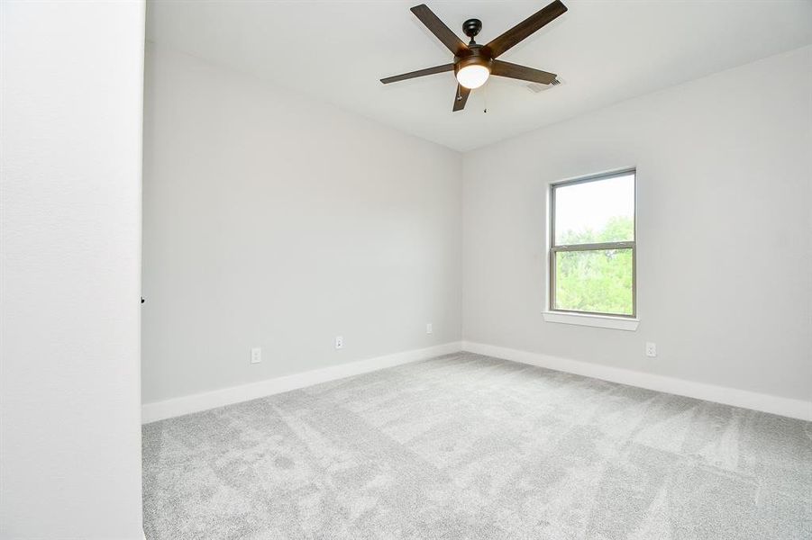 This is a bright, 4th bedroom featuring light gray walls, a textured gray carpet, and a ceiling fan with a light fixture. It has a single window providing natural light and a view of greenery.