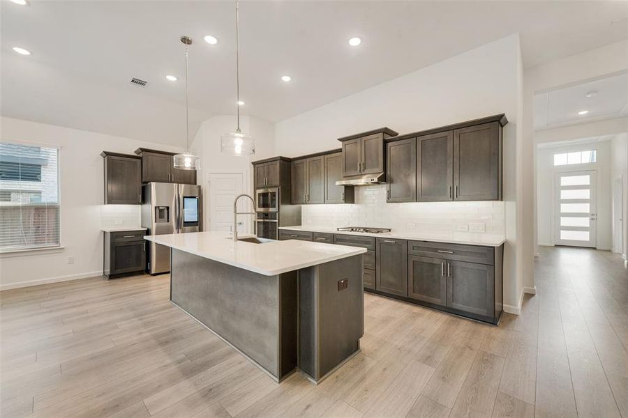 Kitchen featuring decorative light fixtures, backsplash, an island with sink, stainless steel appliances, and light wood-type flooring