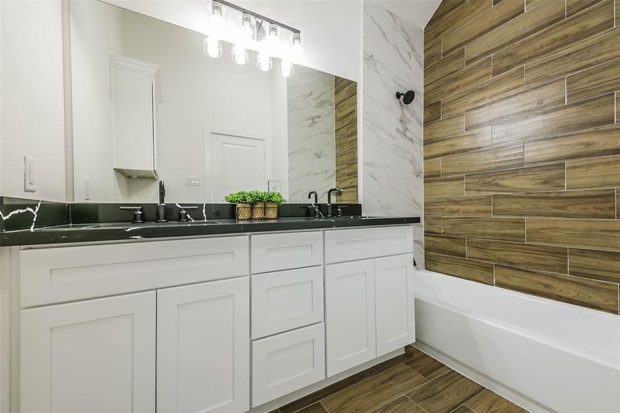 This is a modern bathroom featuring sleek white cabinetry with dark countertops, a large mirror, contemporary lighting, and a mix of marble and wood-textured tiles.