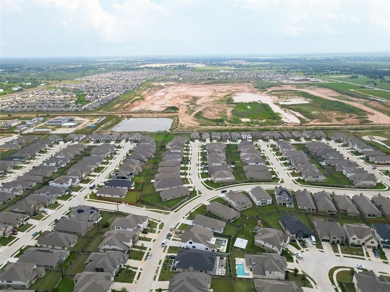 This aerial photograph showcases a suburban residential area with rows of similar, neatly arranged single-family homes. The neighborhood features well-manicured lawns, curving streets, and a new development phase in the background, suggesting potential for community growth. Nearby amenities appear to include a body of water and open green spaces.