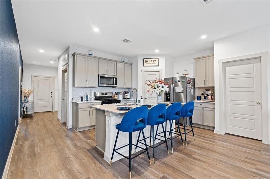 This is a modern, spacious kitchen featuring stainless steel appliances, light gray cabinetry, and a central island with plenty of room for barstools. The room has premium laminate flooring and includes a designated pantry area. Approximate Measurements: 17x11