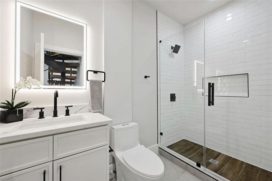 The modern bath on the first floor complements the kitchen's aesthetic with white marble countertops, creating a seamless design theme throughout the home. The shower area is accentuated with wood tile, adding a natural element and a touch of warmth to the sleek and sophisticated bathroom design.