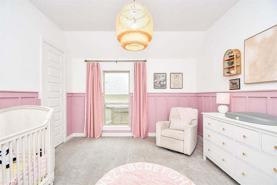 This is a cozy, well-lit nursery room with a white and pastel pink color scheme, featuring wainscoting. It includes a window dressed in pink curtains, providing natural light.