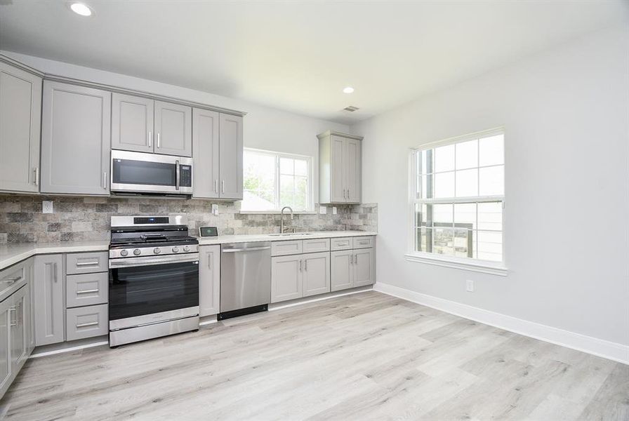 This is a modern kitchen with stainless steel appliances, gray shaker cabinets, stone backsplash, and light wood-look flooring. Large breakfast area It features ample natural light from the large window.