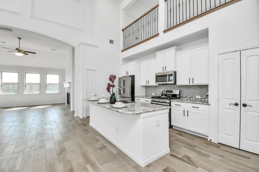 This is a spacious, modern kitchen with high ceilings, white cabinetry, stainless steel appliances, and a large island. The open-concept design flows into the living area and is complemented by ample natural light from multiple windows.