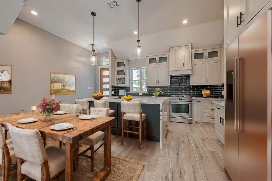 This is a modern, open-concept kitchen and dining space featuring sleek stainless steel appliances, white cabinetry with dark backsplash, a large center island with seating, and a rustic wooden dining table set. The room is well-lit with pendant lights and has light wood flooring.