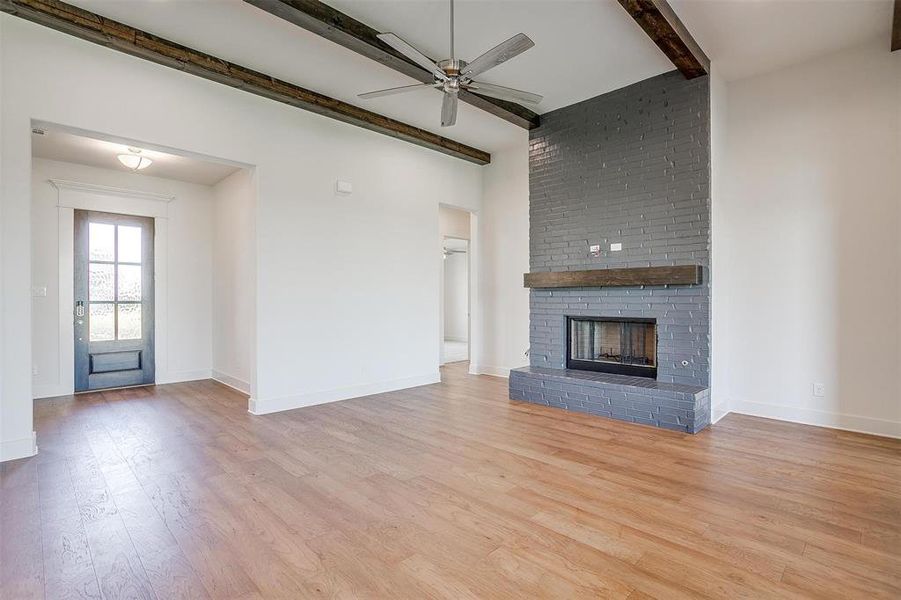 Unfurnished living room with a fireplace, brick wall, light wood-type flooring, beam ceiling, and ceiling fan