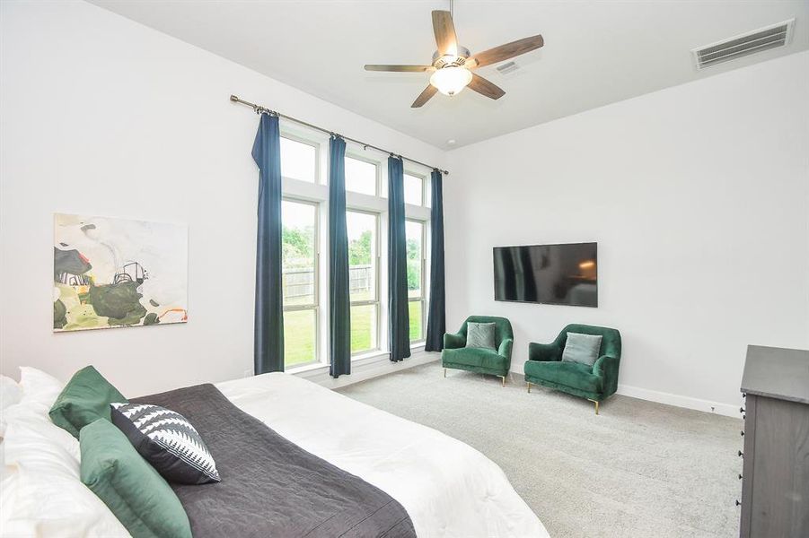 Well-lit primary bedroom featuring neutral walls, newly carpeted flooring, a ceiling fan, and large windows with a view of greenery outside. Also, includes a cozy sitting area for relaxation.