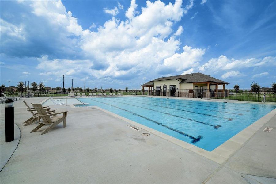 This is a community pool area featuring a large swimming pool with clear blue water and lounge chairs, adjacent to a clubhouse, all enclosed by a fence for privacy and safety. The  well-maintained facility offers a comfortable and inviting outdoor space for relaxation and recreation.