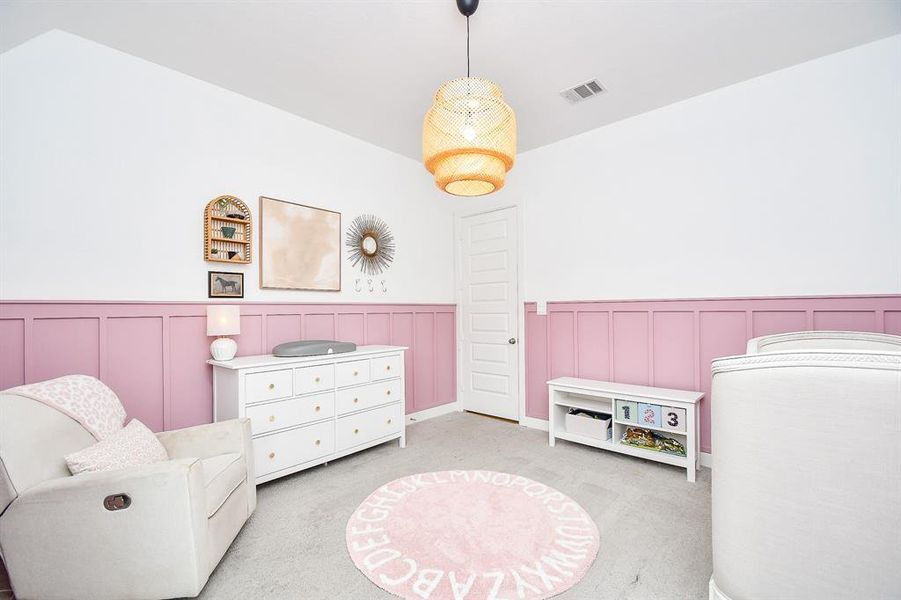 This is a cozy, well-lit nursery room with a white and pastel pink color scheme, featuring wainscoting. It includes a window dressed in pink curtains, providing natural light.