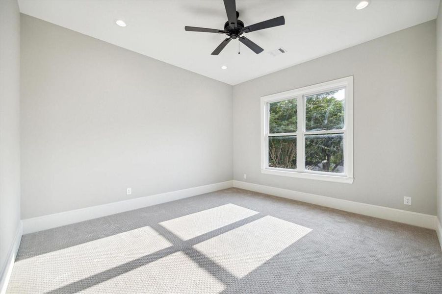 This is a bright, empty room with neutral walls, a large window providing natural light, and a ceiling fan. It has a carpeted floor with a small area rug placed in the center.