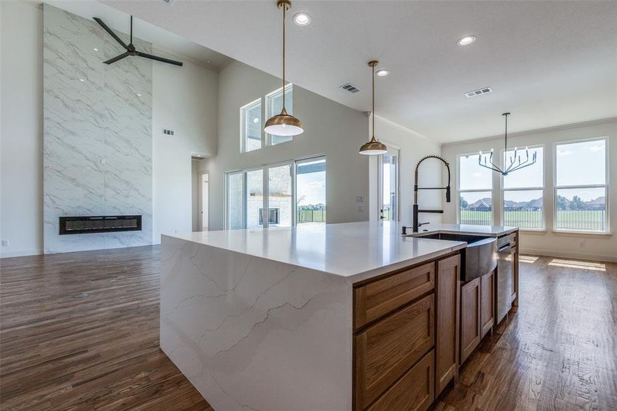 Kitchen featuring dark wood-type flooring, an island with sink, ceiling fan with notable chandelier, pendant lighting, and a premium fireplace