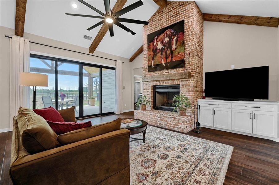 Living room with beam ceiling, dark wood-type flooring, a brick fireplace, high vaulted ceiling, and ceiling fan