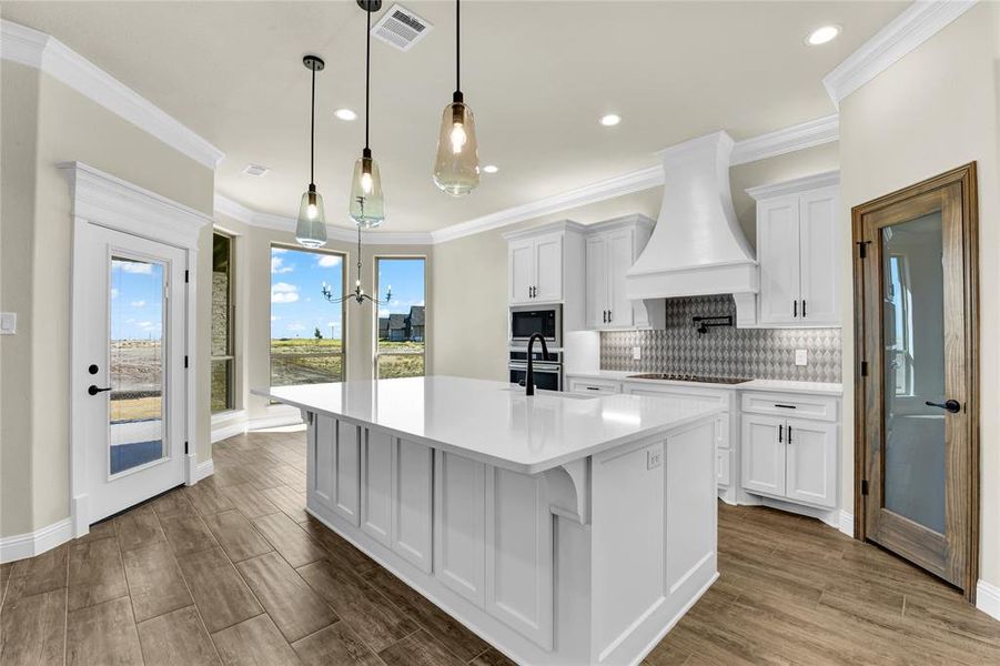 Kitchen with a kitchen island with sink, custom range hood, wood-type flooring, white cabinetry, and appliances with stainless steel finishes