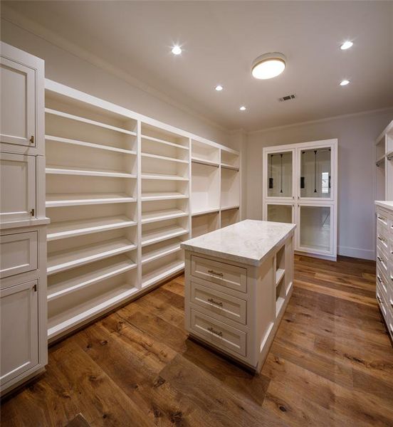 Ladies' spacious dressing room with extensive custom built-ins, pull down hanging rods, center island, deep storage cabinets, hampers, a concealed safe and engineered French Oak wood floors