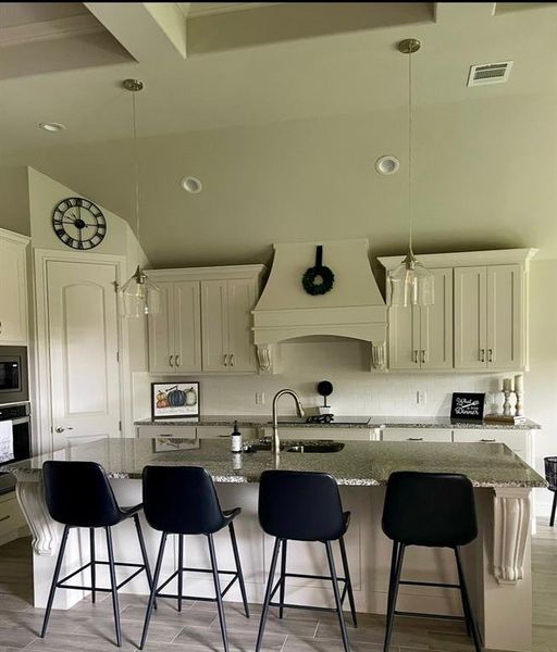 Kitchen featuring an island with sink, stone countertops, hanging light fixtures, custom range hood, and light hardwood / wood-style flooring