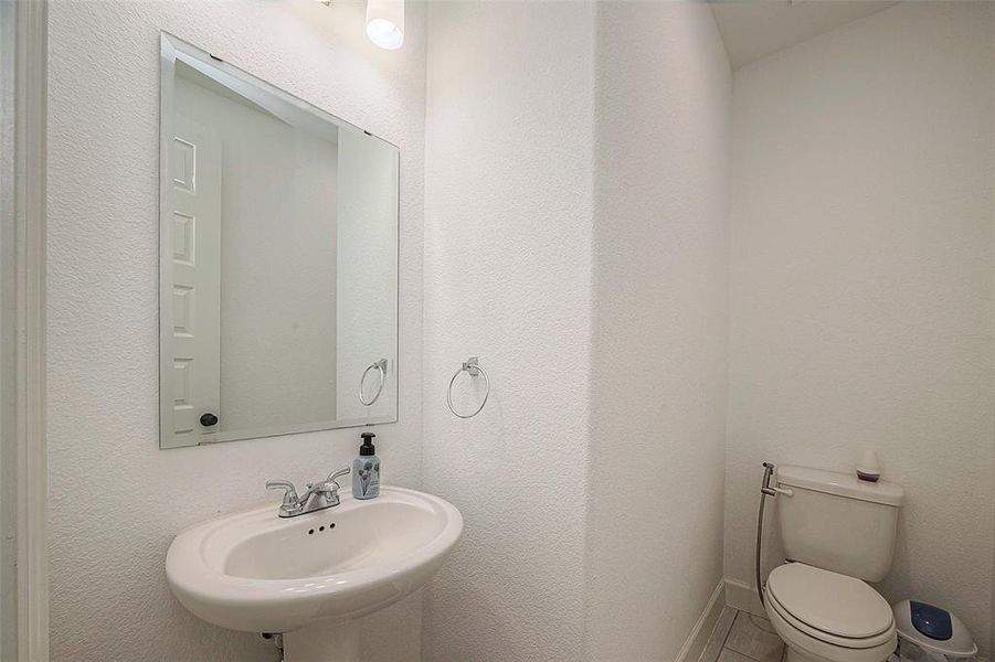 This is a compact half-bathroom featuring a pedestal sink, a toilet, a large mirror, and overhead lighting. The walls are painted white, giving the space a clean and bright appearance.