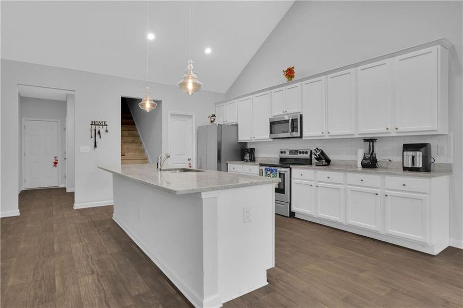 Kitchen with dark hardwood / wood-style flooring, a center island with sink, appliances with stainless steel finishes, and decorative light fixtures