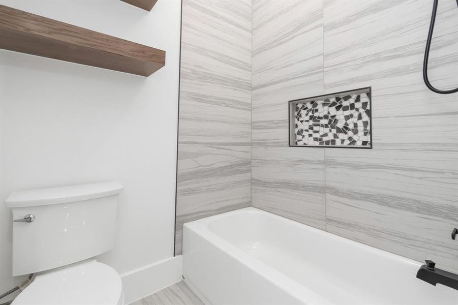 This is a modern bathroom featuring a white bathtub with a black faucet, a white toilet, and light gray marble-like wall tiles. There's a unique mosaic tile accent in the shower area and a wooden shelf for storage.