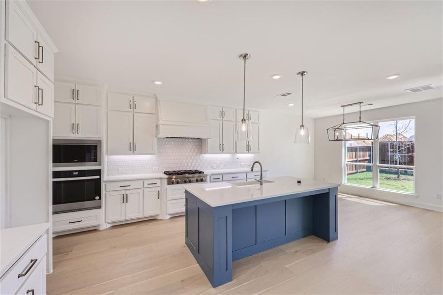 Kitchen featuring light hardwood / wood-style flooring, appliances with stainless steel finishes, a kitchen island with sink, backsplash, and custom exhaust hood