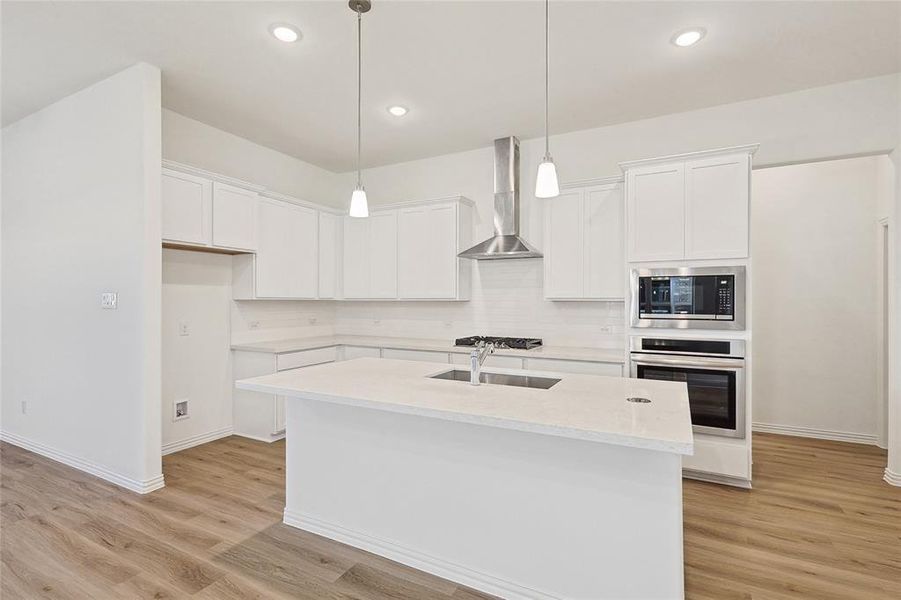 Kitchen with white cabinets, hanging light fixtures, a kitchen island with sink, wall chimney range hood, and stainless steel appliances