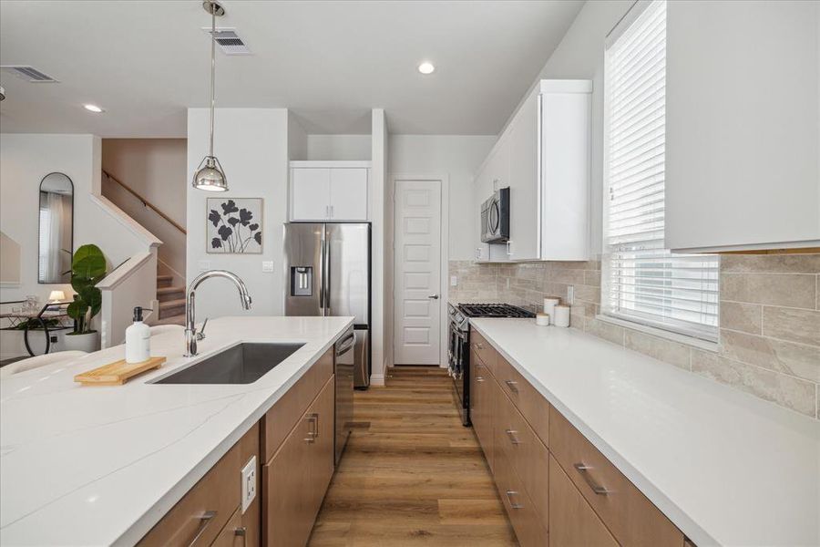 This sleek kitchen layout features a spacious quartz island with a modern stainless steel sink, perfectly complementing the stainless steel appliances. Ample storage is provided by the natural wood-tone cabinetry, while the neutral tile backsplash adds warmth and texture to the bright, open space.