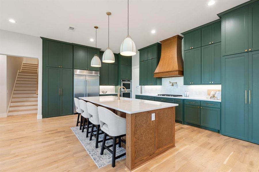 Kitchen with backsplash, hanging light fixtures, custom exhaust hood, an island with sink, and light wood-type flooring