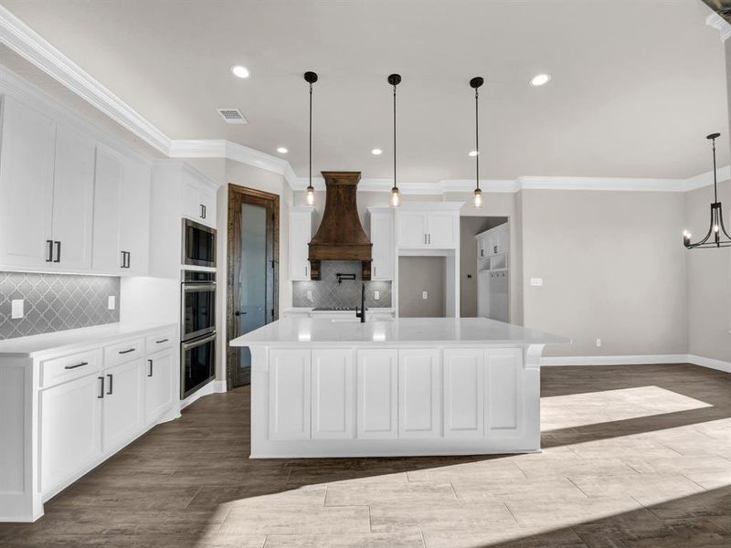 Kitchen with stainless steel appliances, pendant lighting, custom range hood, backsplash, and a kitchen island with sink