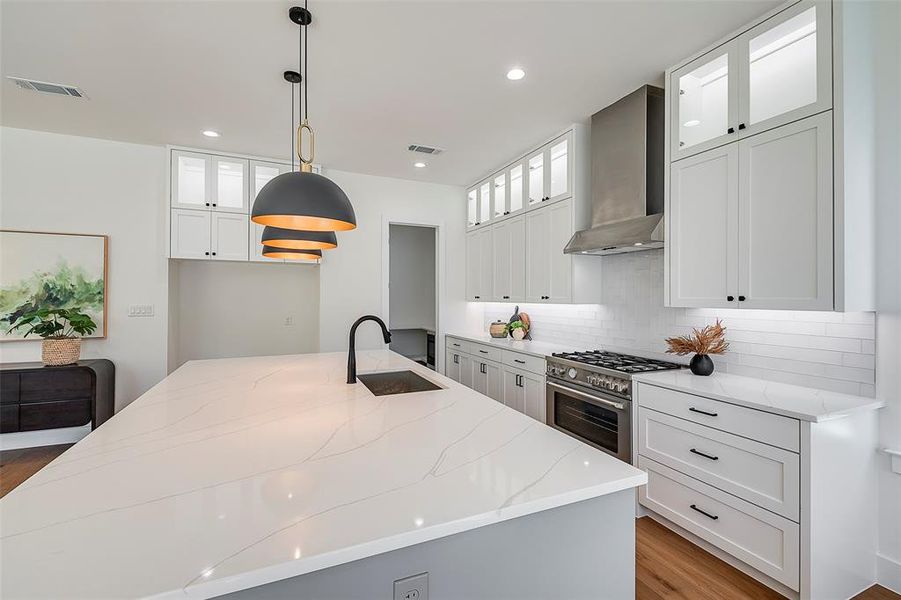 Kitchen featuring wall chimney exhaust hood, a kitchen island with sink, sink, decorative light fixtures, and white cabinets