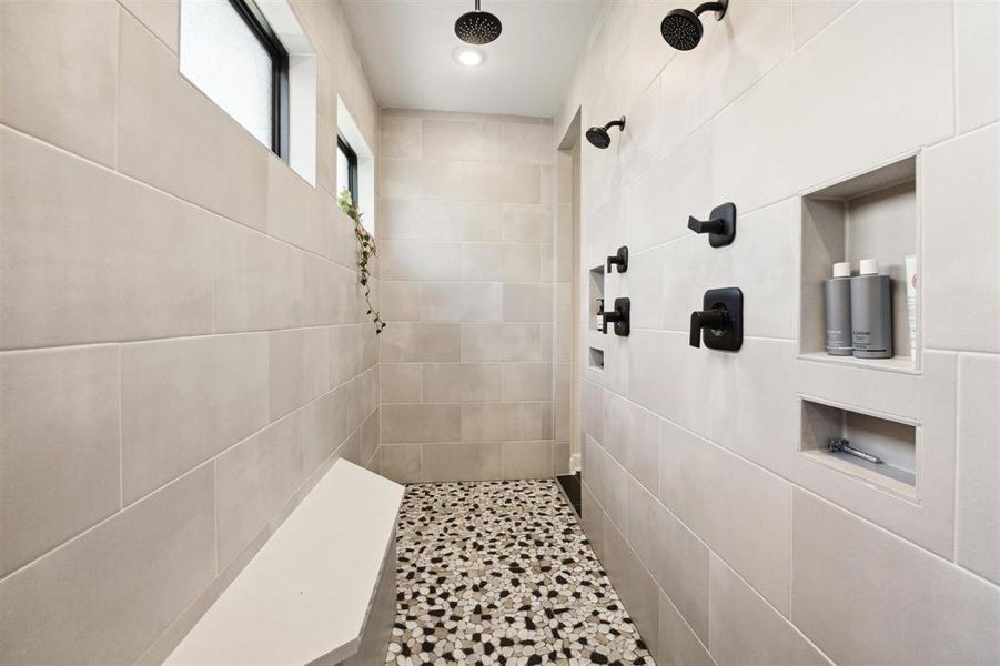 You'll love the huge walk-in shower featuring large beige tiles, dual showerheads, built-in shelving for toiletries, and a stylish pebble-tiled floor. There's also a bench for added comfort, and a small window allows for natural light while maintaining privacy.