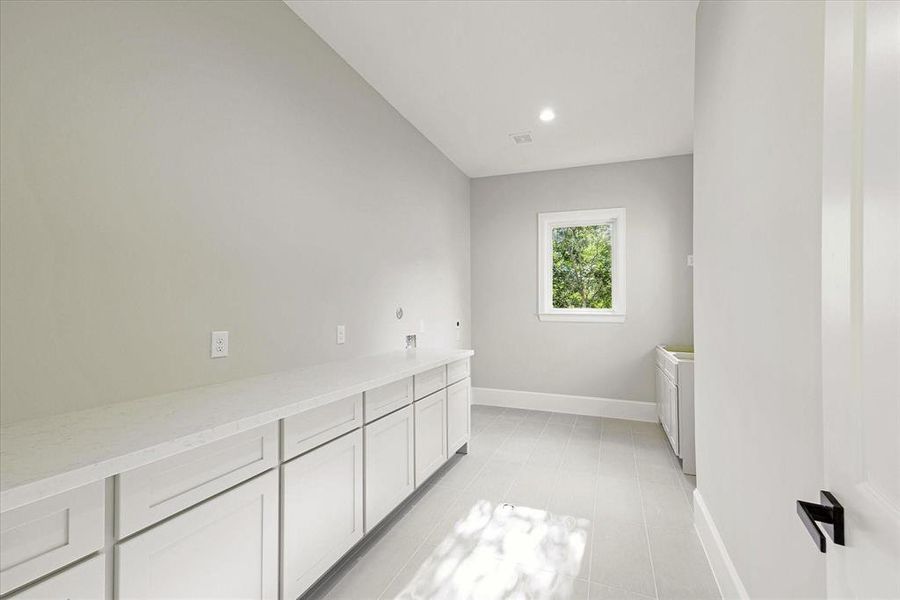 The photo shows a bright, modern laundry room with ample white cabinetry, sleek countertops, and a single window that lets in natural light. The room features tile flooring and recessed lighting.