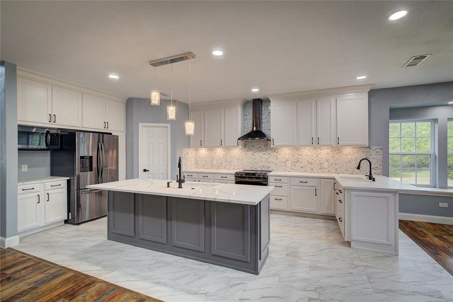 Kitchen with wall chimney style range hood, stainless steel appliances, hanging light fixtures, light wood-type flooring, and a center island with sink