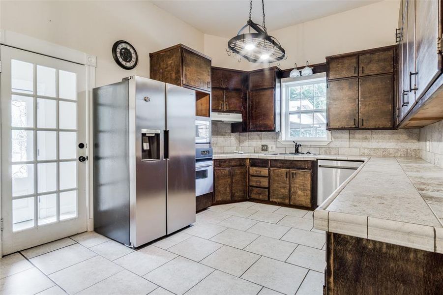 Kitchen with light tile patterned floors, backsplash, stainless steel appliances, hanging light fixtures, and dark brown cabinetry