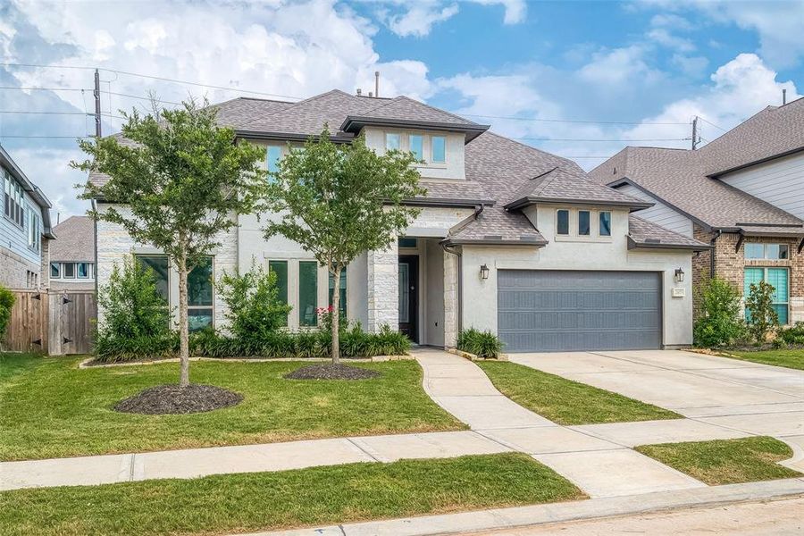 This is a modern two-story home featuring a stone and stucco facade with a well-manicured lawn and two young trees in the front yard. The residence includes an attached two-car garage and boasts a contemporary design in a suburban setting.