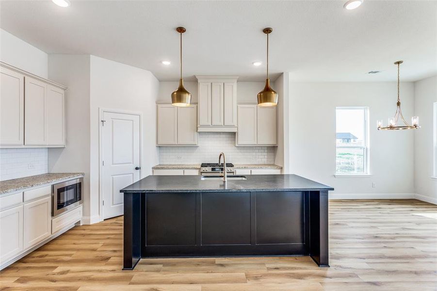 Kitchen with light hardwood / wood-style floors, backsplash, black microwave, an island with sink, and pendant lighting