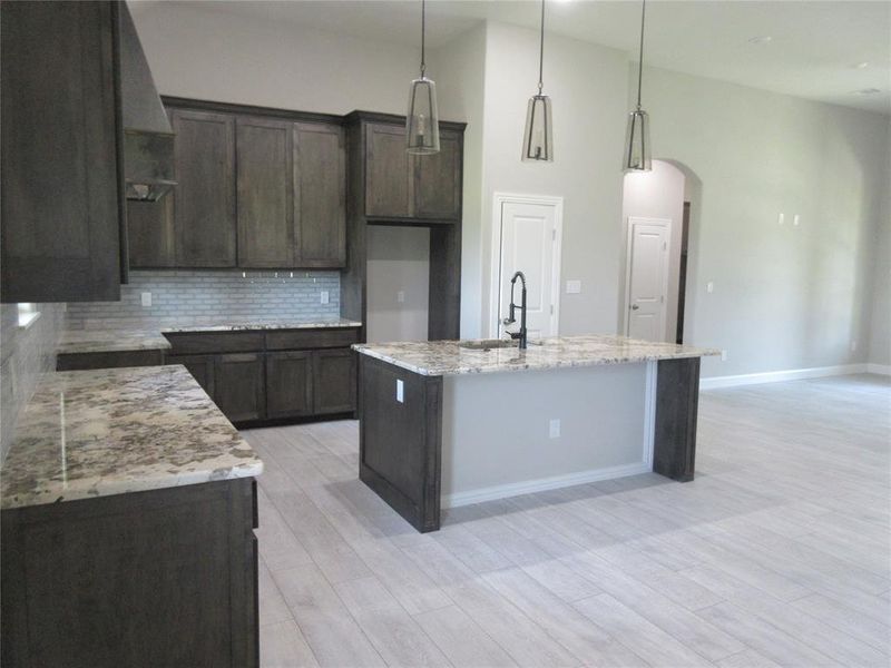 Kitchen with dark brown cabinets, a center island with sink, backsplash, light stone counters, and sink