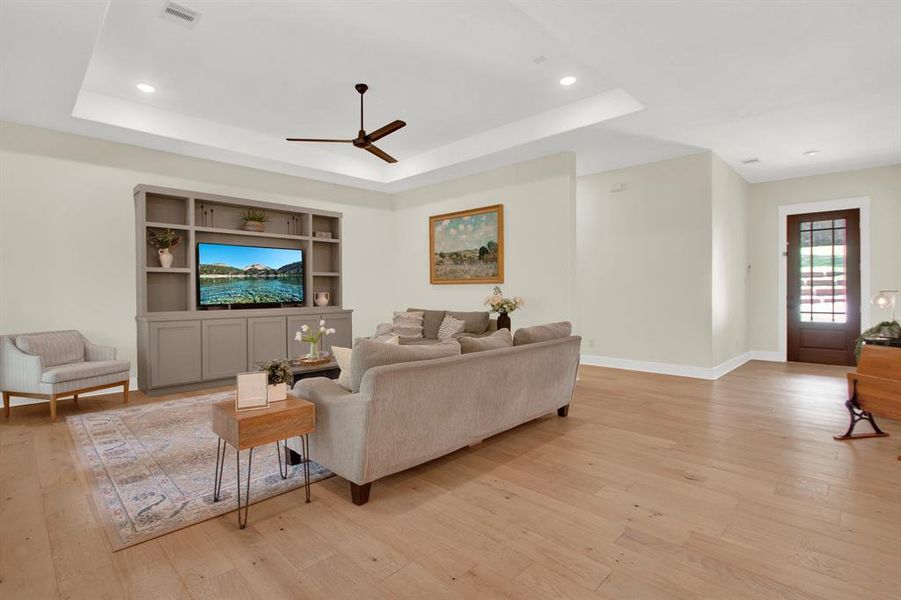 This is a spacious and well-lit living room featuring a neutral color palette, hardwood floors, a built-in entertainment unit with shelving, and a modern ceiling fan. A glass-paneled door provides natural light and access to the exterior.