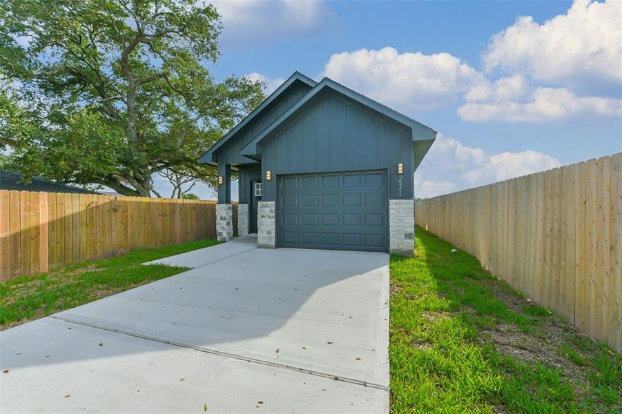 This is a modern single-story home featuring a sleek gray exterior with stone column accents. It includes an attached garage and is surrounded by a new wooden fence, providing privacy. A mature tree in the front yard adds to the curb appeal.