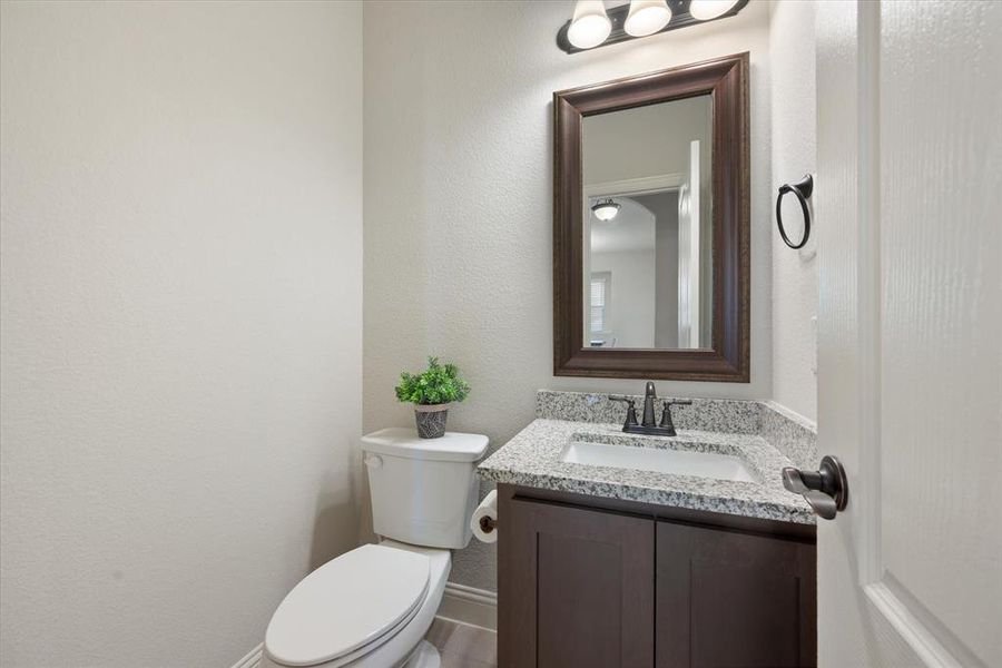 Guest half bath downstairs features granite top vanity, framed mirror, bronze fixtures and hardware, engineered wood flooring and elongated toilet.