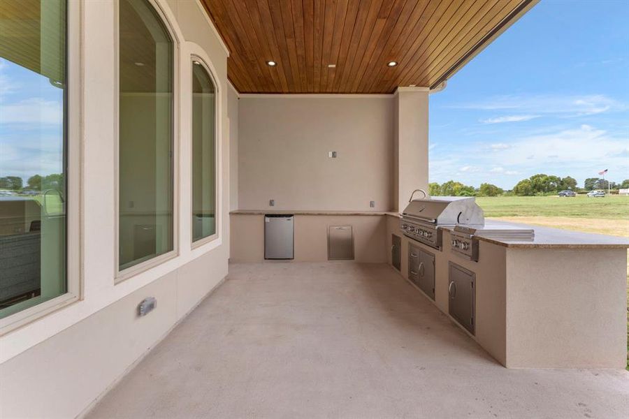 This outdoor kitchen features a built-in barbecue grill, fridge, and counter space, perfect for outdoor dining and entertaining. Notice the sleek wood planked ceiling that adds warmth to the space and large windows that likely provide a view and natural light from the home's interior.