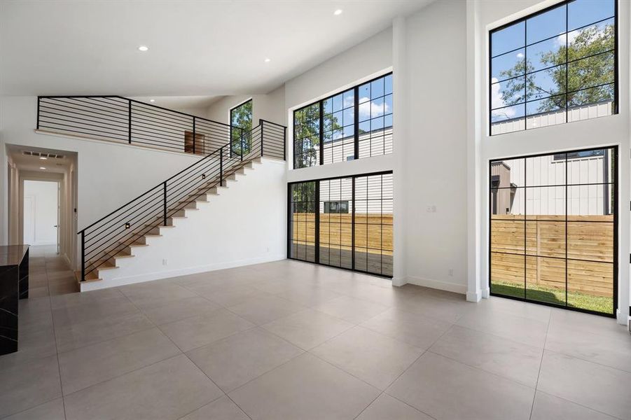 This is a spacious, modern living area featuring high ceilings, large windows for ample natural light, a sleek staircase with a contemporary railing design, and tile flooring. It offers a view of the backyard through floor-to-ceiling glass doors.