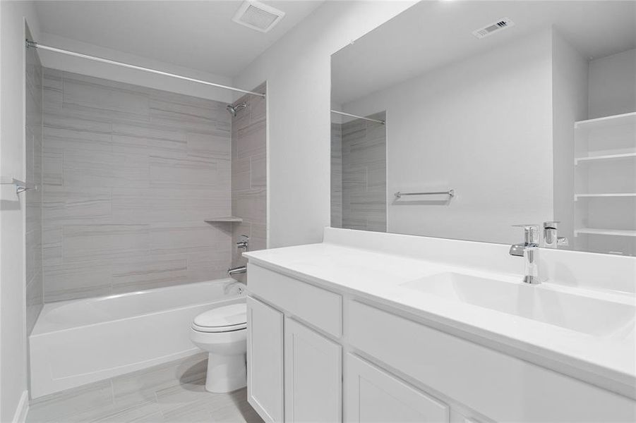 Secondary bath features tile flooring, bath/shower combo with tile surround, white stained wood cabinets, beautiful light countertops, mirror, dark, sleek fixtures and modern finishes.