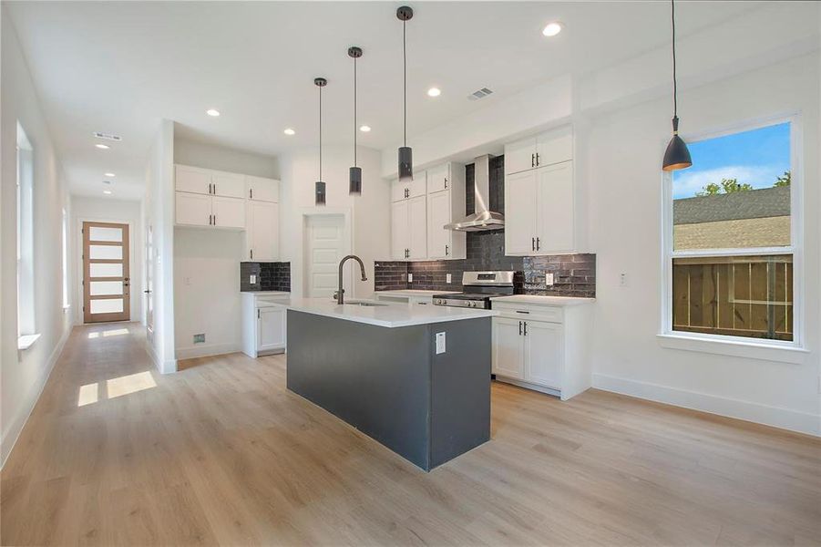 Kitchen with an island with sink, wall chimney exhaust hood, hanging light fixtures, and stainless steel stove