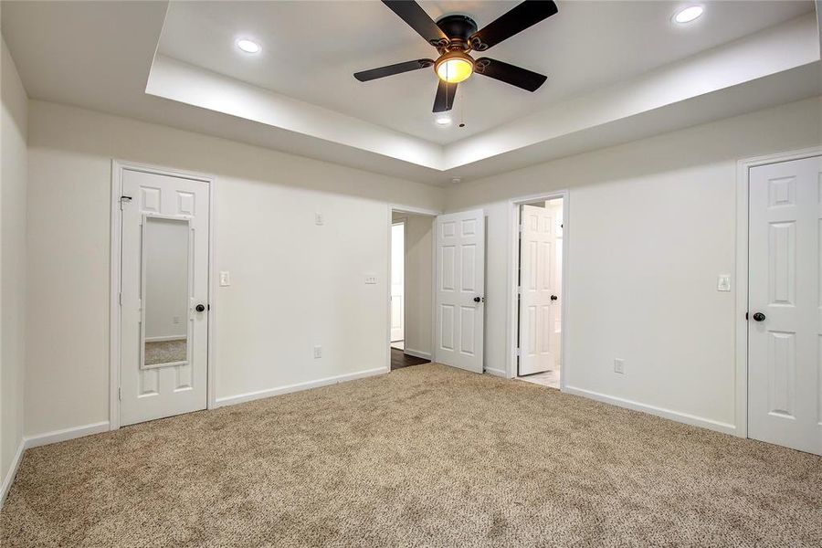 Primary bedroom with a tray ceiling, ensuite bath, ceiling fan, double closets, and carpet floors
