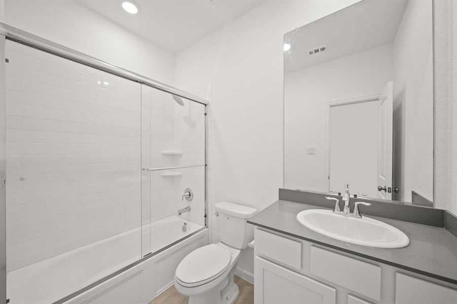 This is a designer bathroom featuring a combination shower/tub with a glass door, a white vanity with a sink and a large mirror, quartz countertop, alongside a standard toilet. The room is finished with white tiles and neutral tones, creating a bright and airy space.