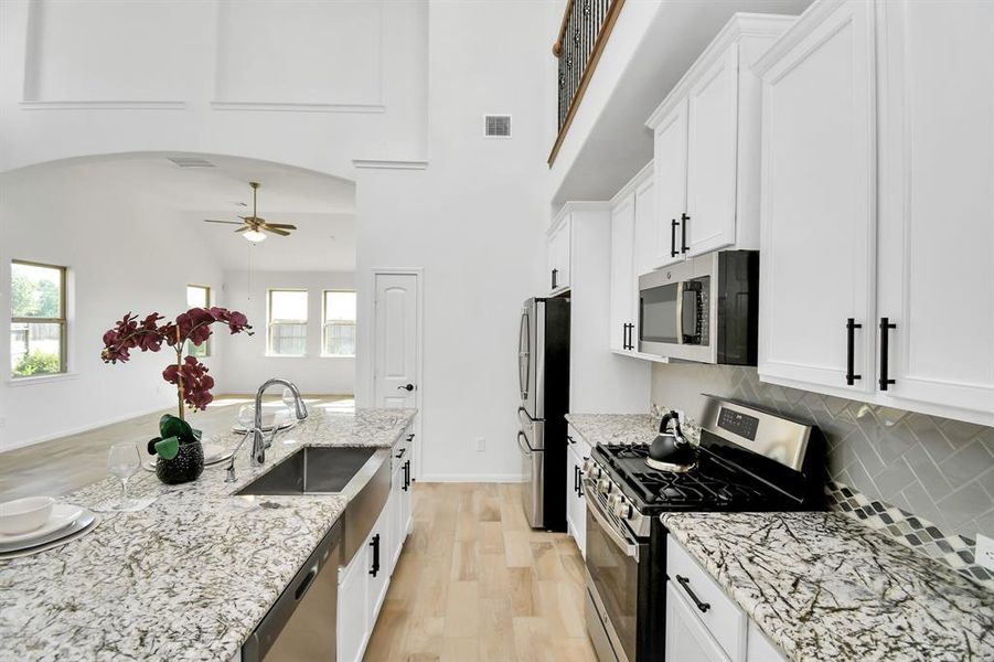 This is a modern, bright kitchen featuring white cabinetry, stainless steel appliances, granite countertops, and a herringbone tile backsplash. The space opens to a high-ceiling living area with ample natural light.