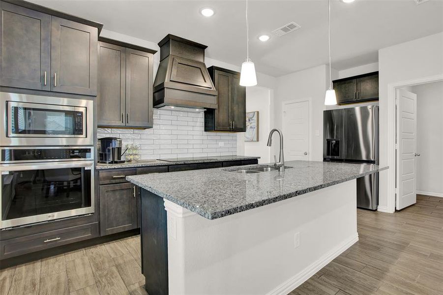 Kitchen with tasteful backsplash, stainless steel appliances, a center island with sink, hanging light fixtures, and custom range hood
