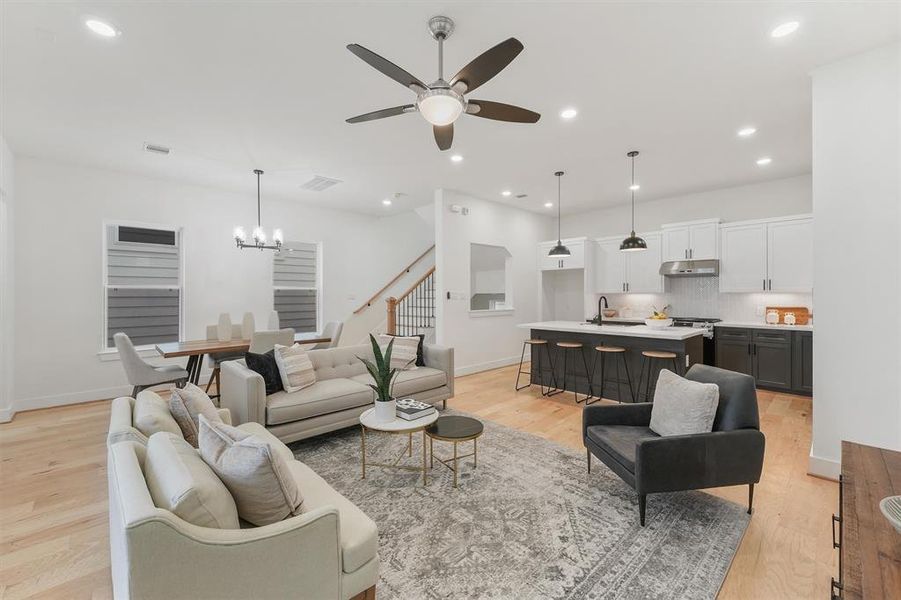 Modern open-plan living space featuring a combined living room and kitchen, with engineered hardwood floors, custom paint, contemporary lighting, and a staircase leading to your 3rd level. The kitchen includes white cabinetry, a central island with seating, and stainless steel appliances.