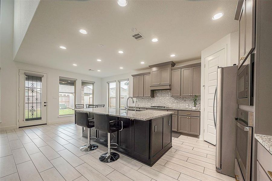 This is a modern kitchen with stainless steel appliances, dark cabinetry, granite countertops, and a central island with bar seating. It features recessed lighting, tile flooring, and has a glass door leading to the outside, providing ample natural light.